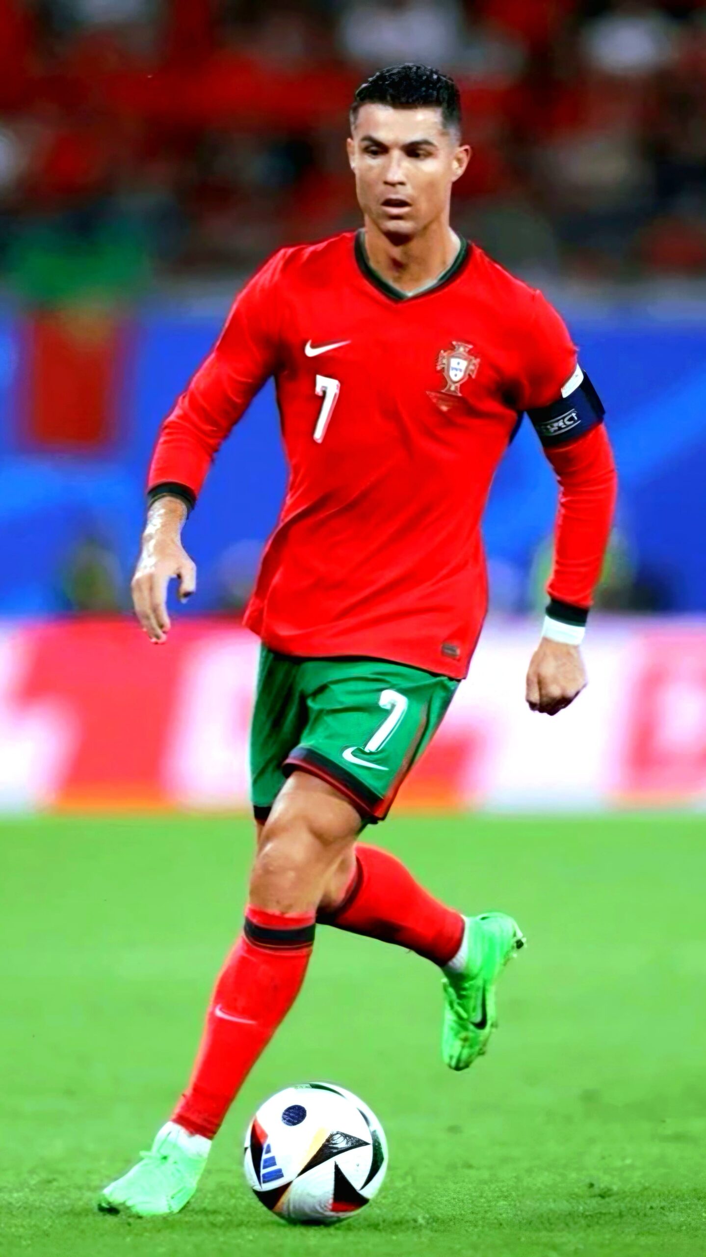 A soccer player in a red and green uniform with the number 7 dribbles a ball on the field, wearing green cleats and a captain’s armband. The background is blurred with blue and red colors, capturing the intensity of the match as seen on IPTV BrasiL.