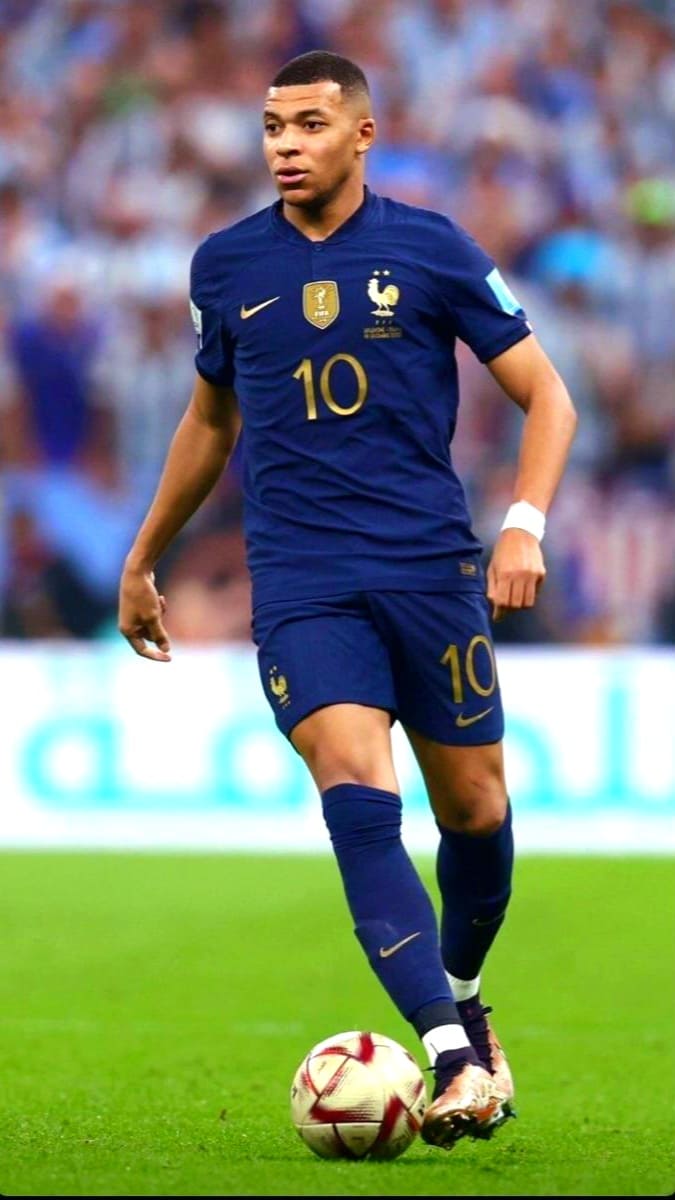 A French soccer player in a blue jersey with number 10 is dribbling a ball on a field during a match, as fans who follow the game live through Brasil IPTV cheer from the blurred background of spectators.